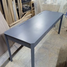 Fabryka Mebli Farmahei - Długi, szary stół z dwoma otworami na kable, w warsztacie stolarskim z widocznym drewnem i narzędziami w tle.