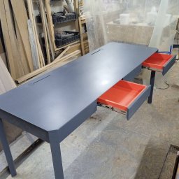 Fabryka Mebli Farmahei - Długi, szary stół biurowy z dwoma wysuniętymi, pomarańczowymi szufladami, widoczny w warsztacie stolarskim z narzędziami i drewnem w tle.