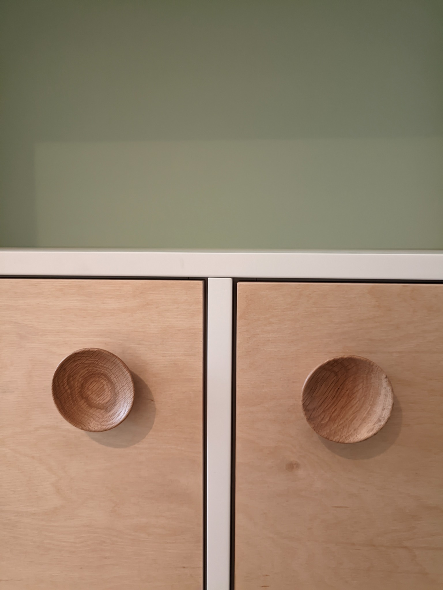 Dwa fronty szuflad z jasnej sklejki, wyposażone w okrągłe, drewniane uchwyty w kształcie miseczek, umieszczone na tle zielonej ściany.