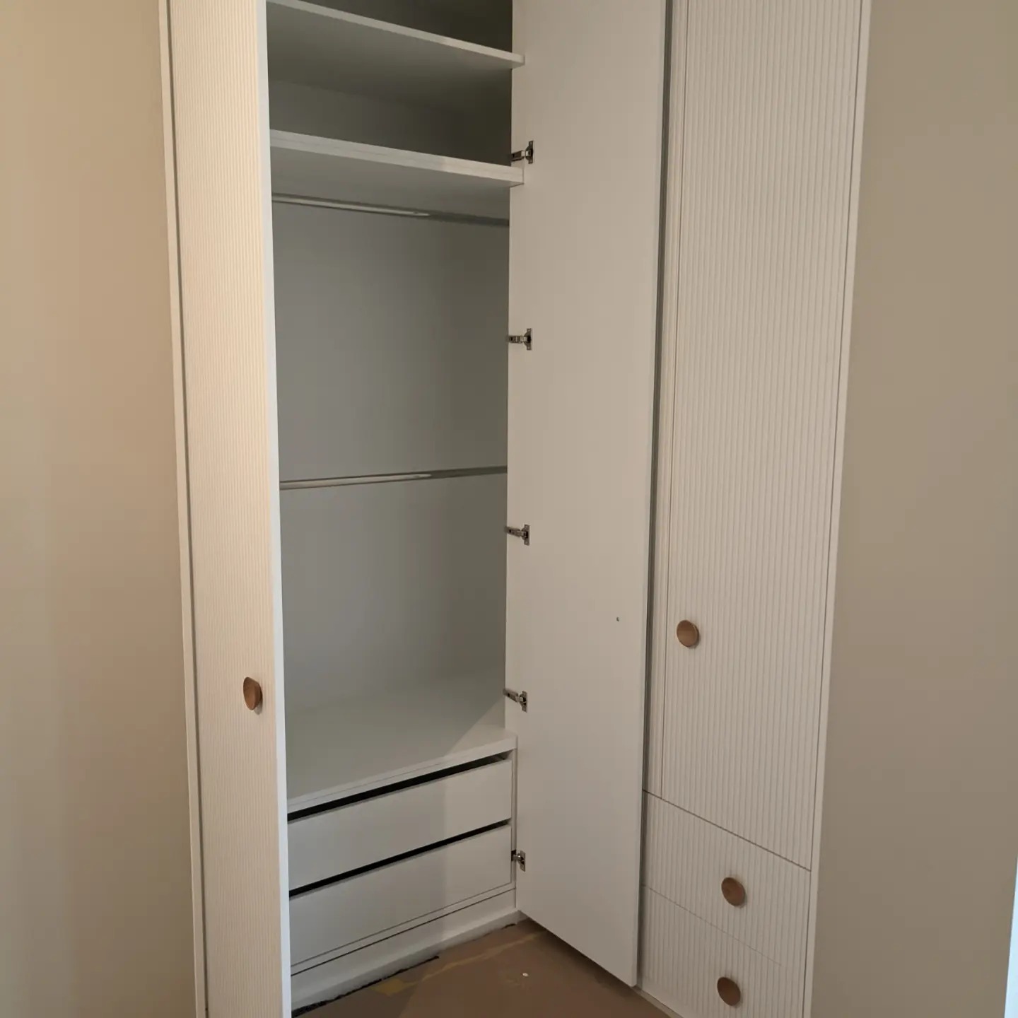 Wnętrze nowoczesnej, białej szafy z ryflowanymi drzwiami, widoczne półki, drążek na ubrania i dwie szuflady, otwarte drzwi ukazują zawartość.