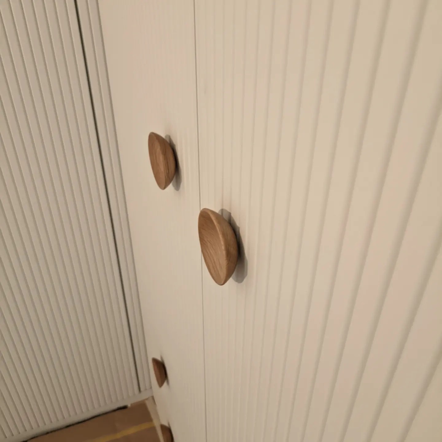 Biała szafa z frezowanymi drzwiami i drewnianymi, półokrągłymi uchwytami, widoczne z bliska pod kątem.