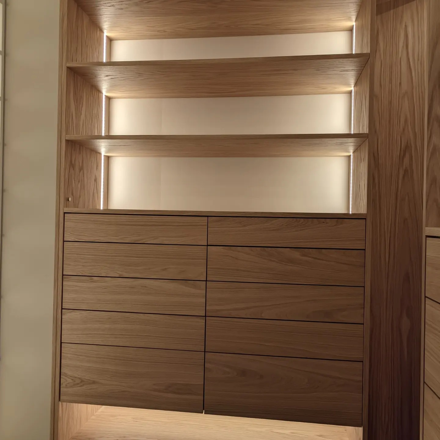 Drewniana szafa na wymiar z oświetleniem LED, widoczne półki i szuflady z minimalistycznymi uchwytami, ciepłe światło podkreśla strukturę drewna.