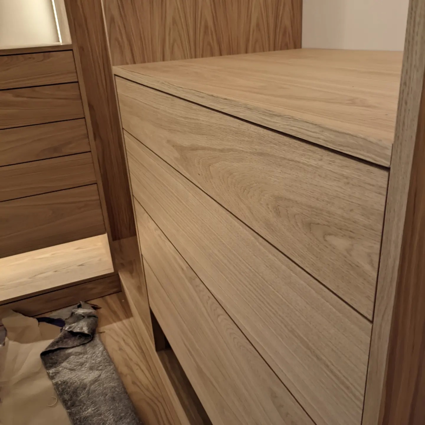Fragment drewnianej szafy z szufladami, widoczna faktura drewna i krawędzie, na podłodze leżą materiały zabezpieczające podczas montażu.