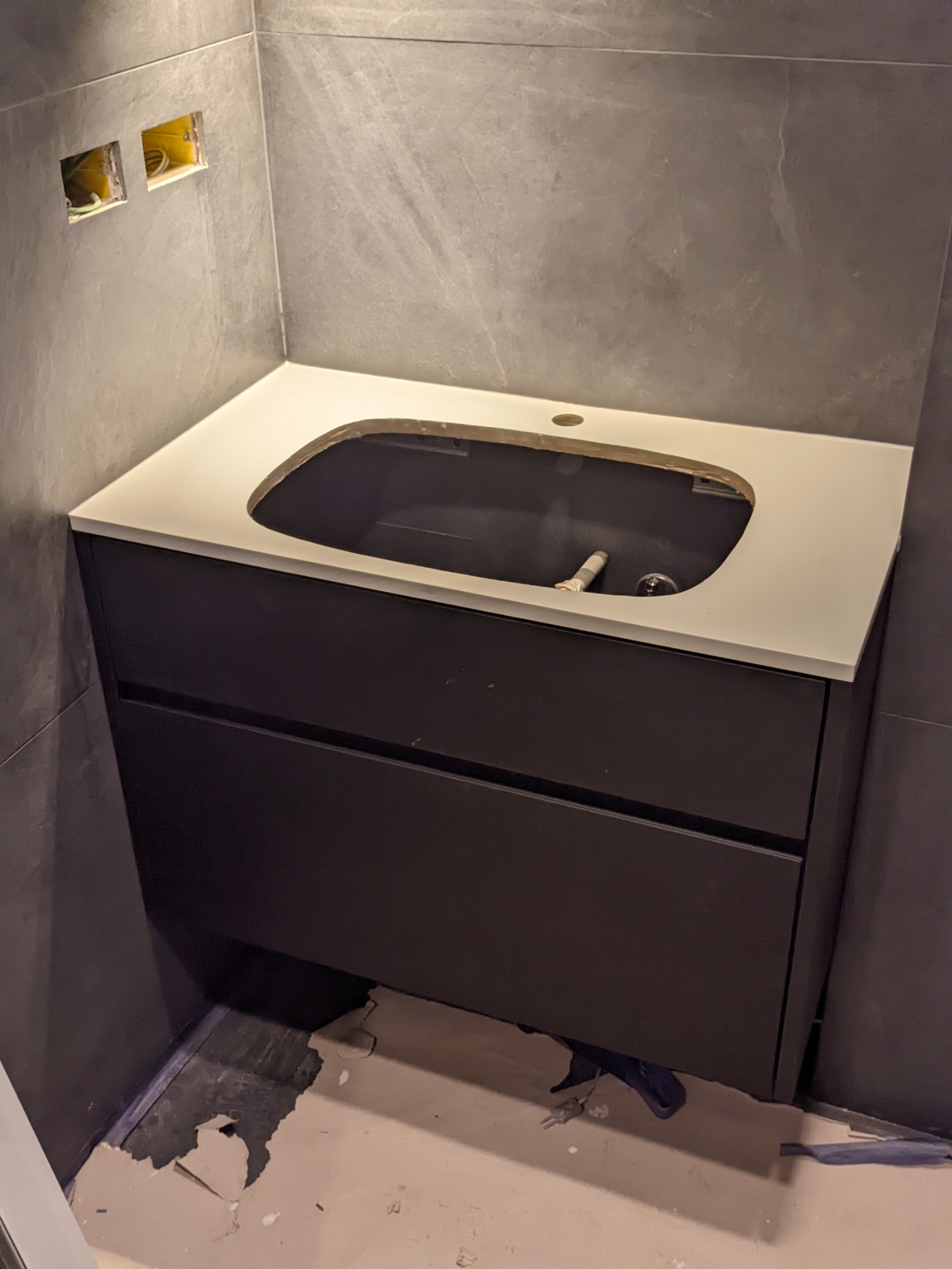 Montaż szafki łazienkowej z jasnym blatem w surowym wnętrzu, widoczne niedokończone prace wykończeniowe i wystające kable elektryczne.