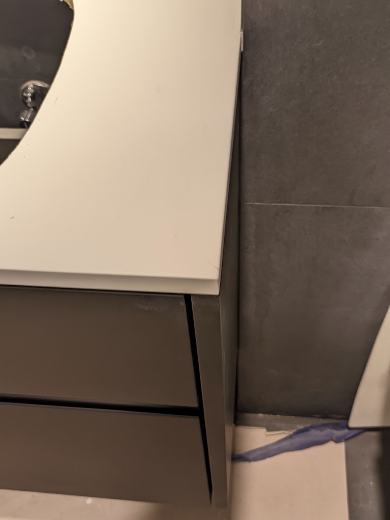 Fragment łazienki z umywalką nablatową na szafce z szufladami, widoczne szare płytki na ścianie i silikon zabezpieczający połączenie szafki ze ścianą.