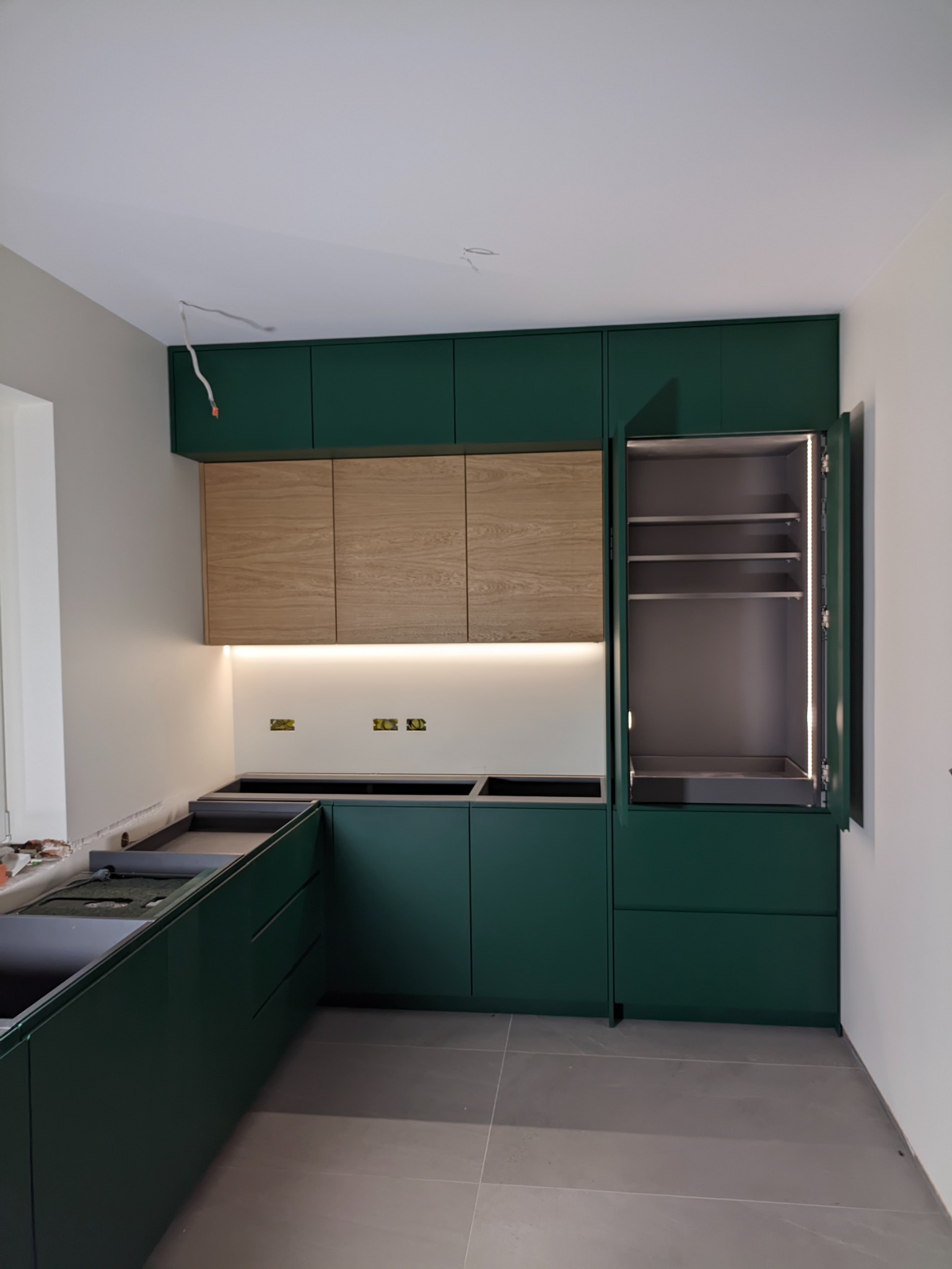 Wnętrze kuchni w trakcie montażu, z zielonymi szafkami dolnymi i górnymi z drewnianymi frontami, podświetleniem LED pod szafkami górnymi oraz otwartą szafką z oświetleniem wewnętrznym. Widoczne...