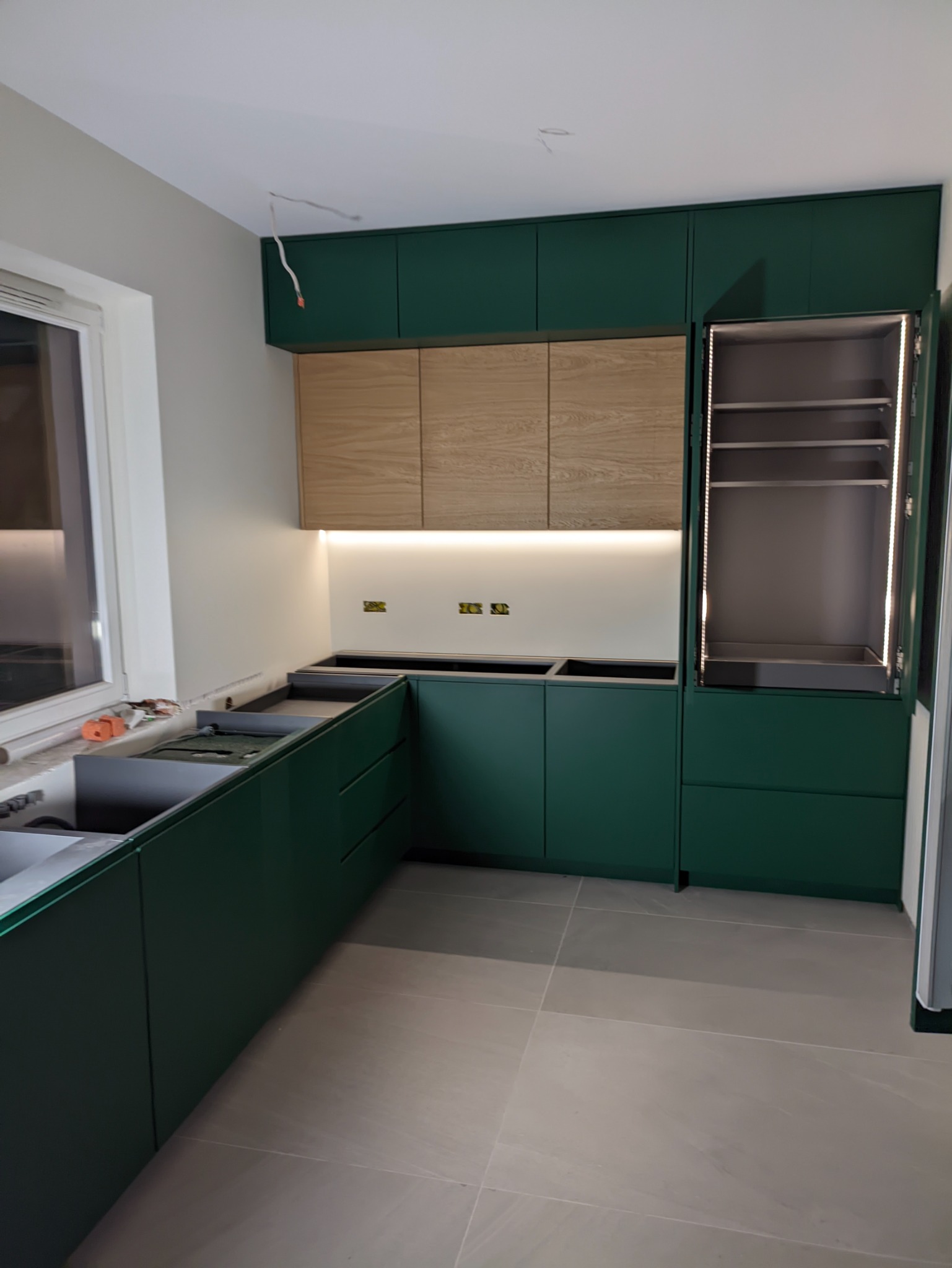 Wnętrze kuchni w trakcie montażu: zielone szafki dolne i górne, drewniane szafki wiszące podświetlone listwą LED, otwarty słupek z oświetleniem wewnętrznym, widoczne instalacje elektryczne, szare...