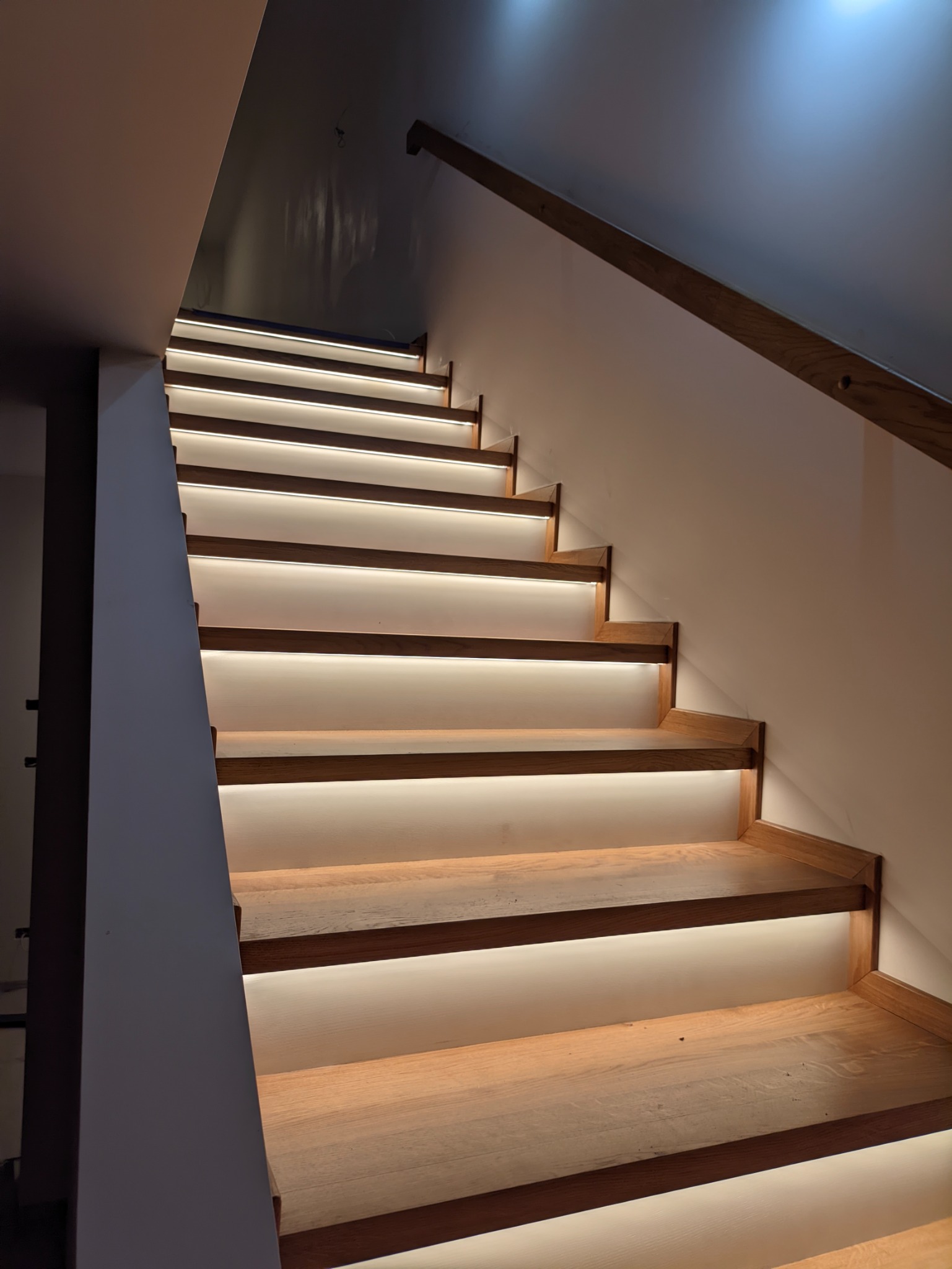 Drewniane schody z podświetleniem LED, widok z dołu, jasne ściany, drewniana poręcz