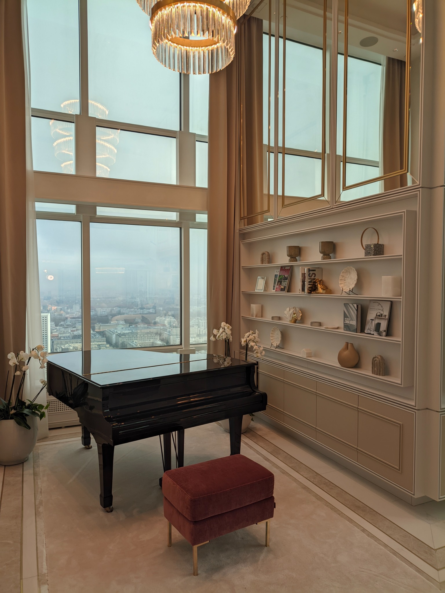 Eleganckie wnętrze z czarnym fortepianem, pufą w kolorze bordo, wysokimi oknami z widokiem na miasto i designerską zabudową meblową z półkami.