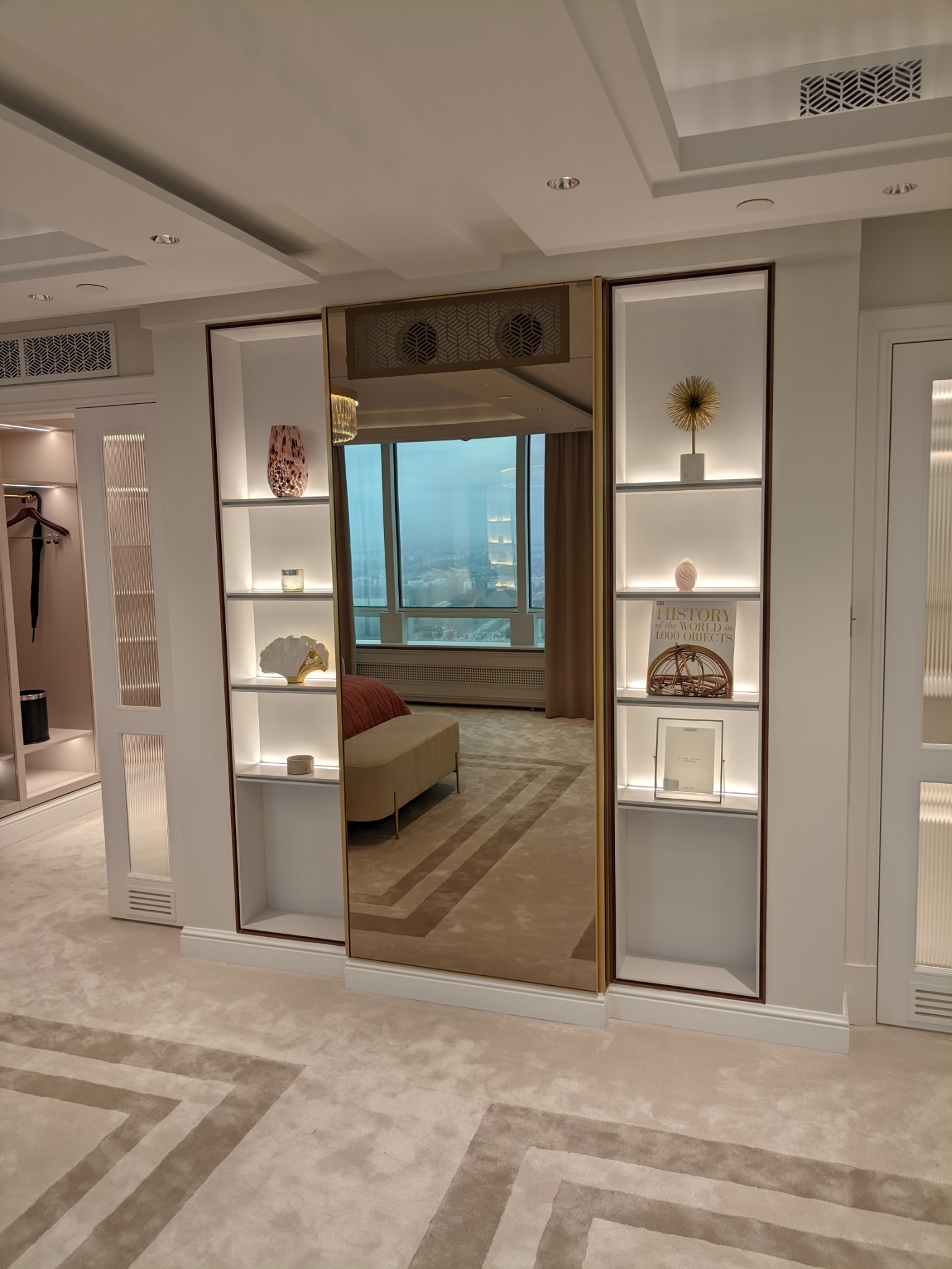 Nowoczesna garderoba z lustrzanymi drzwiami przesuwnymi, oświetlonymi półkami z dekoracjami i widokiem na miasto w tle.