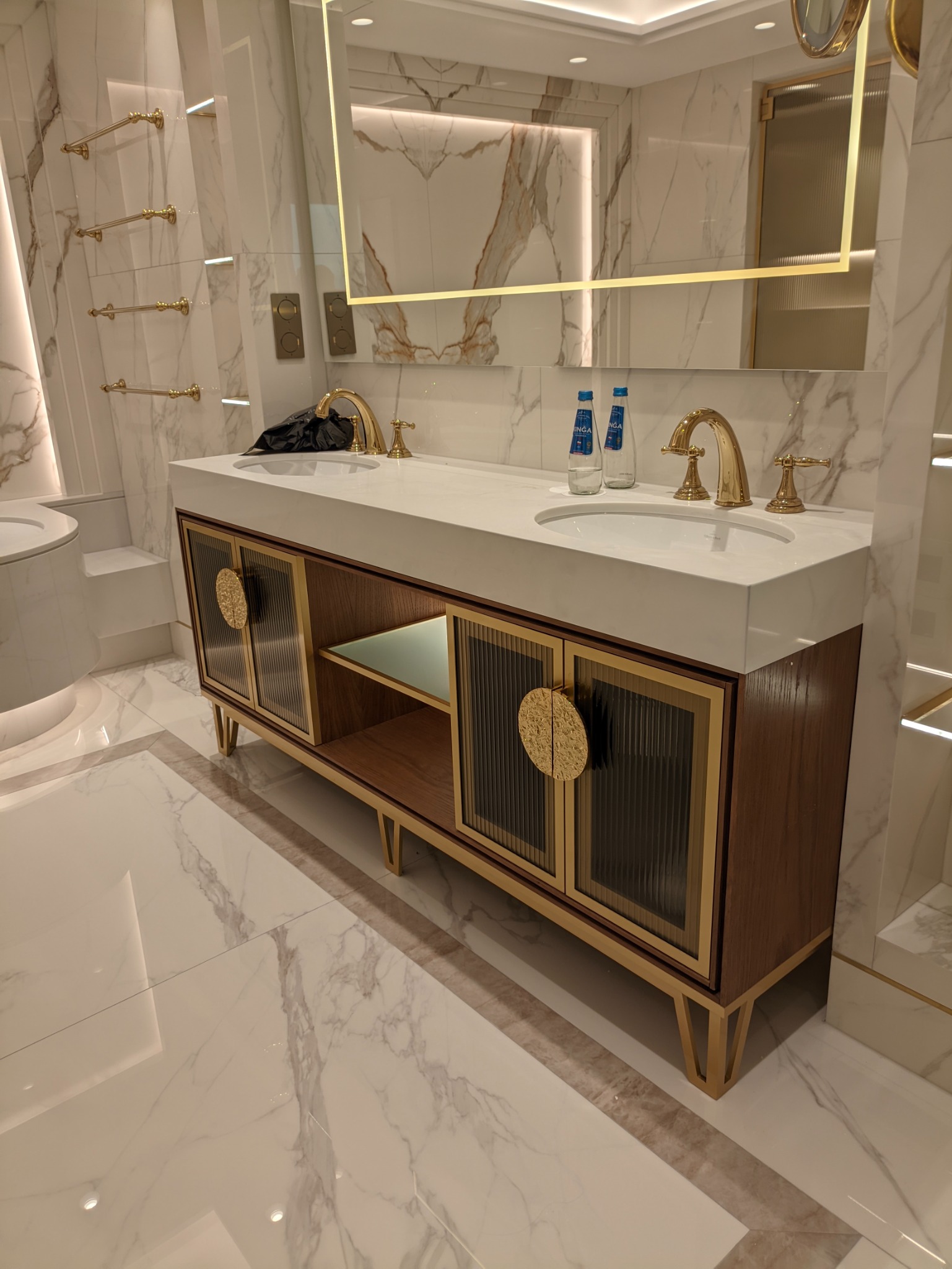 Nowoczesna szafka łazienkowa z dwoma umywalkami, złotymi bateriami i okrągłymi uchwytami na tle marmurowych ścian i podłóg, podświetlane lustro.