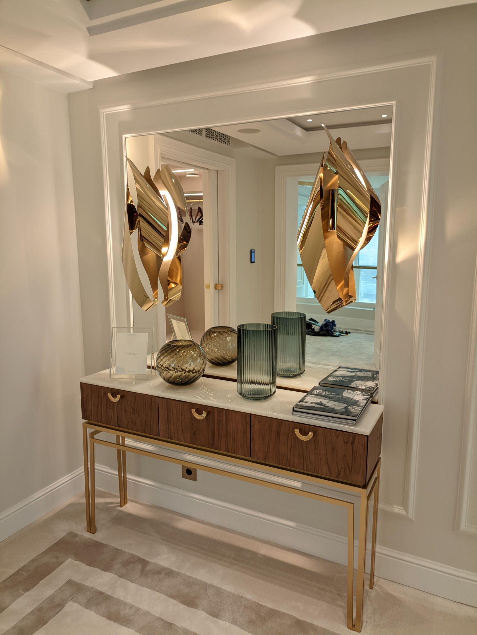 Konsola z drewnianymi szufladami na złotych nogach, biały marmurowy blat, dekoracyjne szkło i książki, odbicie lustrzane wnętrza garderoby z ubraniami.