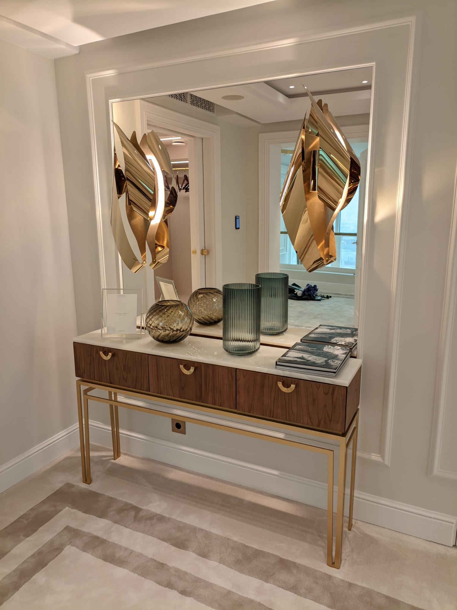 Nowoczesna konsola z drewnianymi szufladami i marmurowym blatem, złote nogi, dekoracje w postaci szklanych wazonów i abstrakcyjnych lamp wiszących, odbicie szafy w lustrze.