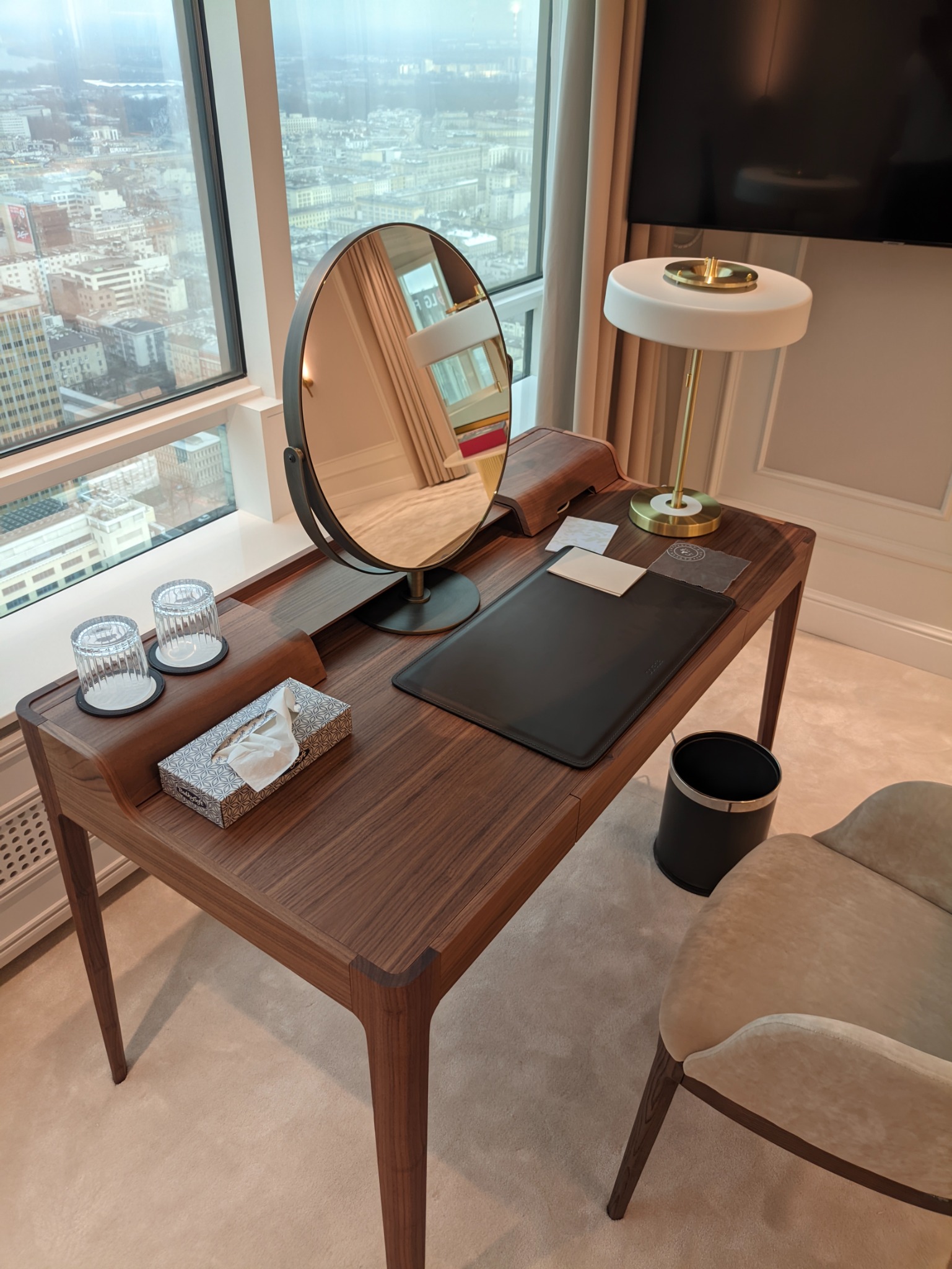 Drewniane biurko z okrągłym lustrem, lampą i akcesoriami biurowymi w pokoju z widokiem na miasto.