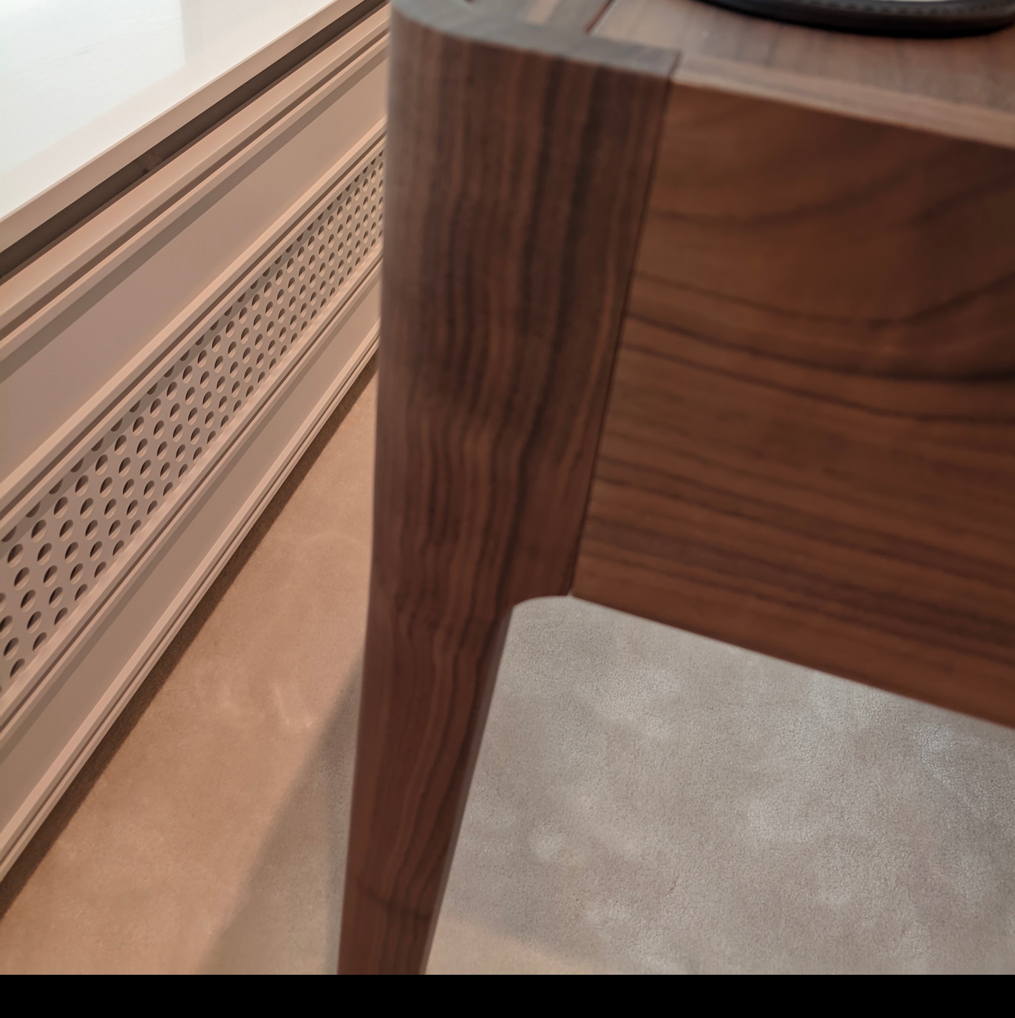 Fragment drewnianego blatu z nogą o zaokrąglonych krawędziach, widoczny na tle beżowej ściany z perforowanym panelem grzejnikowym.