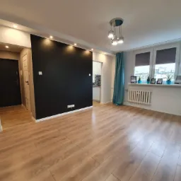 Jasne panele podłogowe w przestronnym salonie z czarną ścianą akcentującą, oświetleniem punktowym i turkusowymi zasłonami, widok na sąsiednie pomieszczenie z wzorzystymi płytkami.