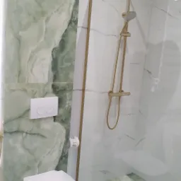 Nowoczesna łazienka z zielonymi marmurkowymi płytkami, złotą armaturą prysznicową i podwieszaną toaletą.