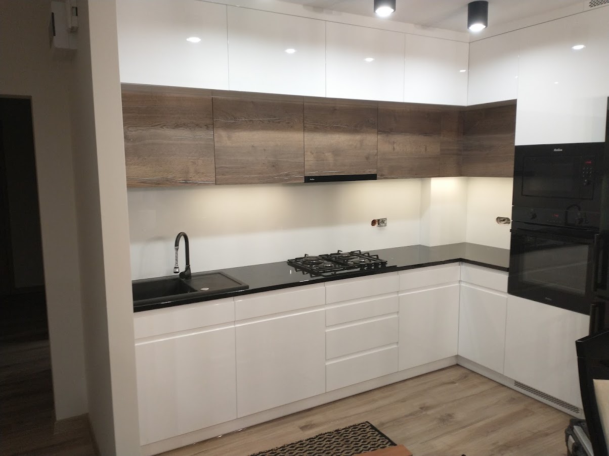 Nowoczesna kuchnia z białymi lakierowanymi frontami i drewnopodobnymi szafkami górnymi w odcieniu dębu halifax cynowanego, czarnym blatem i czarną armaturą, oświetlona punktowymi lampami sufitowymi.