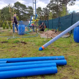 Wiercenie studni na działce: widoczna wiertnica, przygotowane rury w kolorze niebieskim, beczki, dwóch mężczyzn przy pracy, ogrodzenie z siatki w tle.