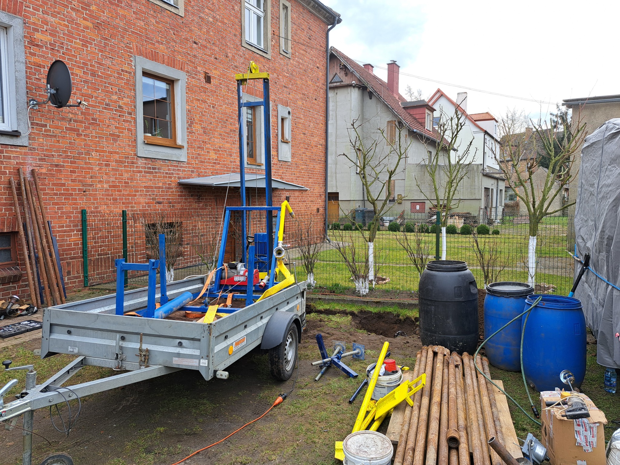 Sprzęt do wiercenia studni głębinowej na przyczepie, z widocznymi rurami wiertniczymi, beczkami na wodę i wykopem w trawie przed ceglanym domem.