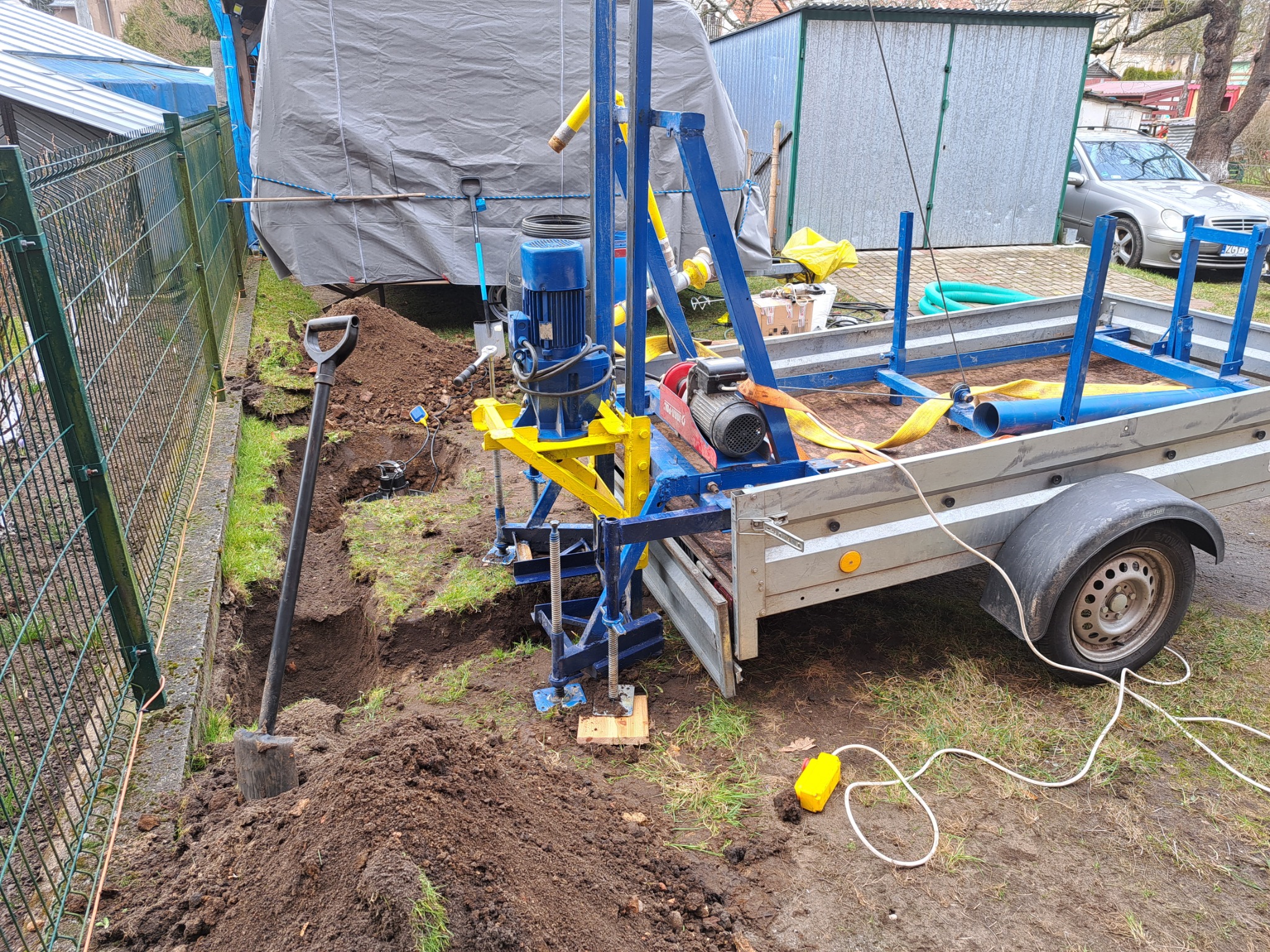 Wiertnica zamontowana na przyczepie obok wykopu z łopatą, teren ogrodzony siatką, w tle budynki gospodarcze i samochód osobowy.