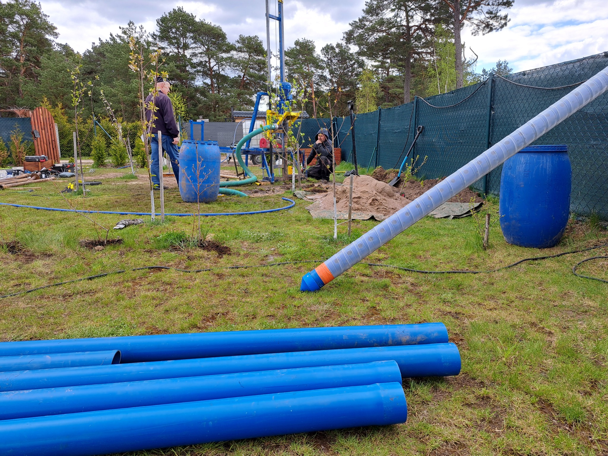 Wiercenie studni na działce: widoczna wiertnica, przygotowane rury w kolorze niebieskim, beczki, dwóch mężczyzn przy pracy, ogrodzenie z siatki w tle.