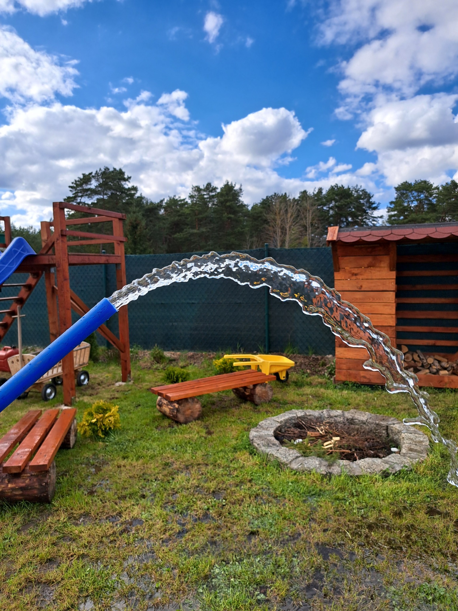 Strumień wody wypływający z niebieskiego węża w kierunku kamiennego paleniska na trawniku, w tle drewniany plac zabaw i szopa na drewno.