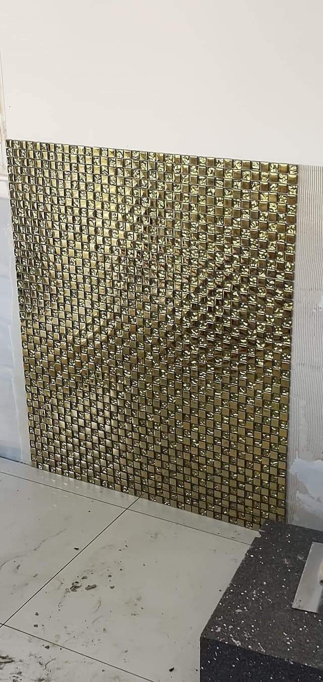 Ściana wyłożona błyszczącą, złotą mozaiką z kwadratowych płytek, widoczne ślady kleju i fugi na białej ścianie obok, fragment marmurowej podłogi.