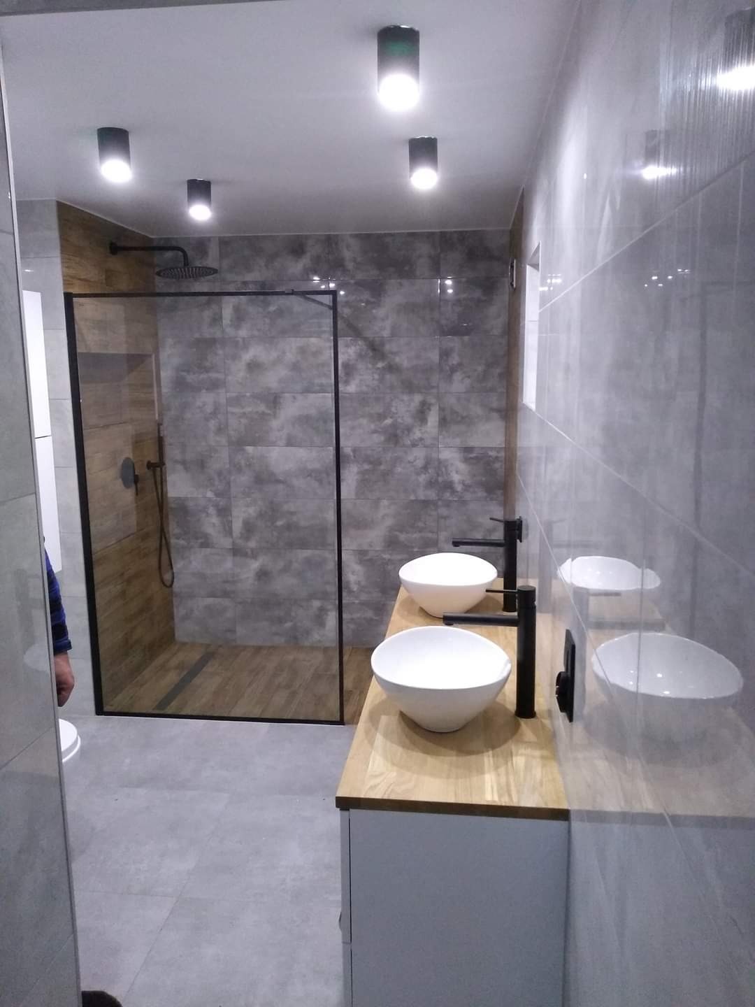 Nowoczesna łazienka z kabiną prysznicową typu walk-in, dwiema okrągłymi umywalkami nablatowymi na drewnianym blacie i szarą glazurą.