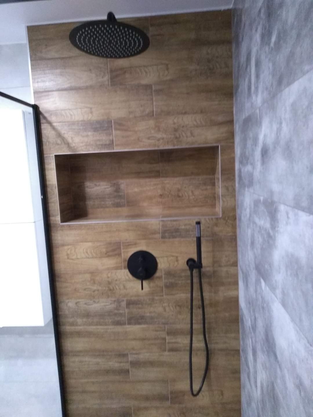 Kabina prysznicowa z deszczownicą i baterią w kolorze czarnym, wykończona płytkami imitującymi drewno oraz szarą mozaiką, z wnęką na kosmetyki.