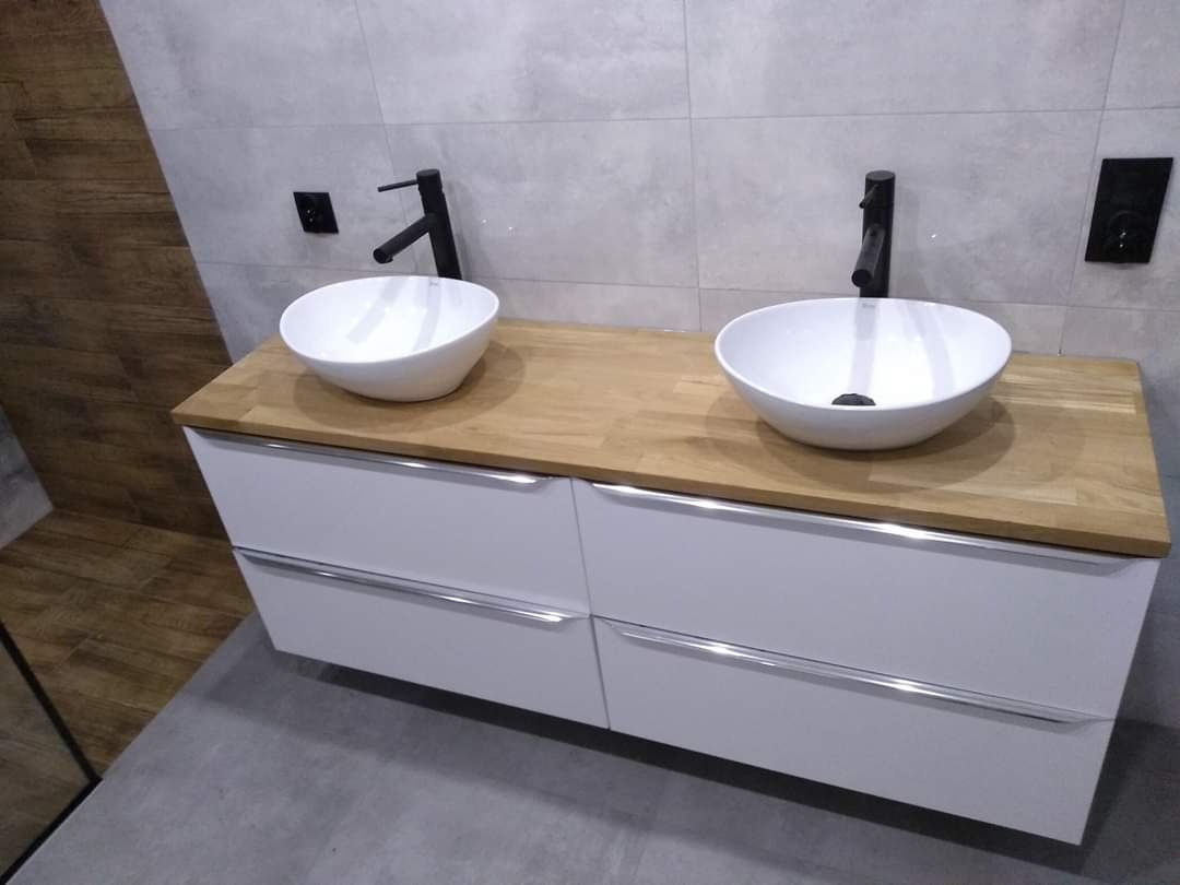 Nowoczesna łazienka z dwiema okrągłymi umywalkami nablatowymi, czarnymi bateriami i blatem z jasnego drewna na białej szafce z szufladami, szare płytki na ścianach i podłodze.