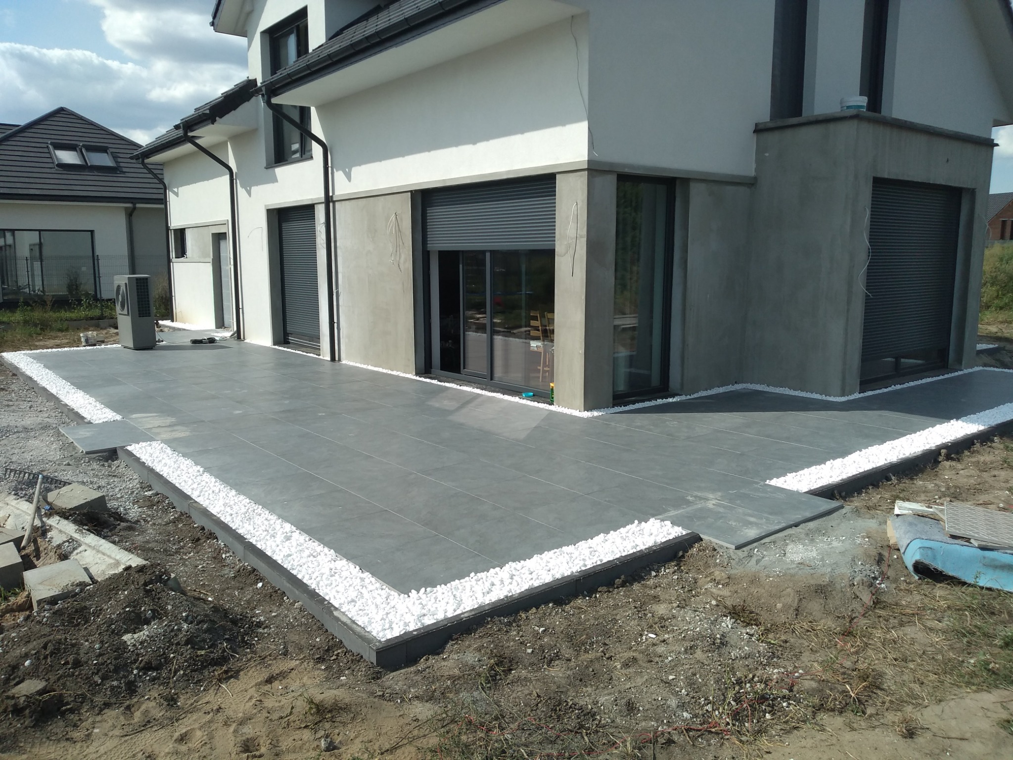 Nowoczesny taras z szarych płyt, obrzeża z białych kamieni, w tle dom z roletami zewnętrznymi, widoczne niedokończone prace ziemne.