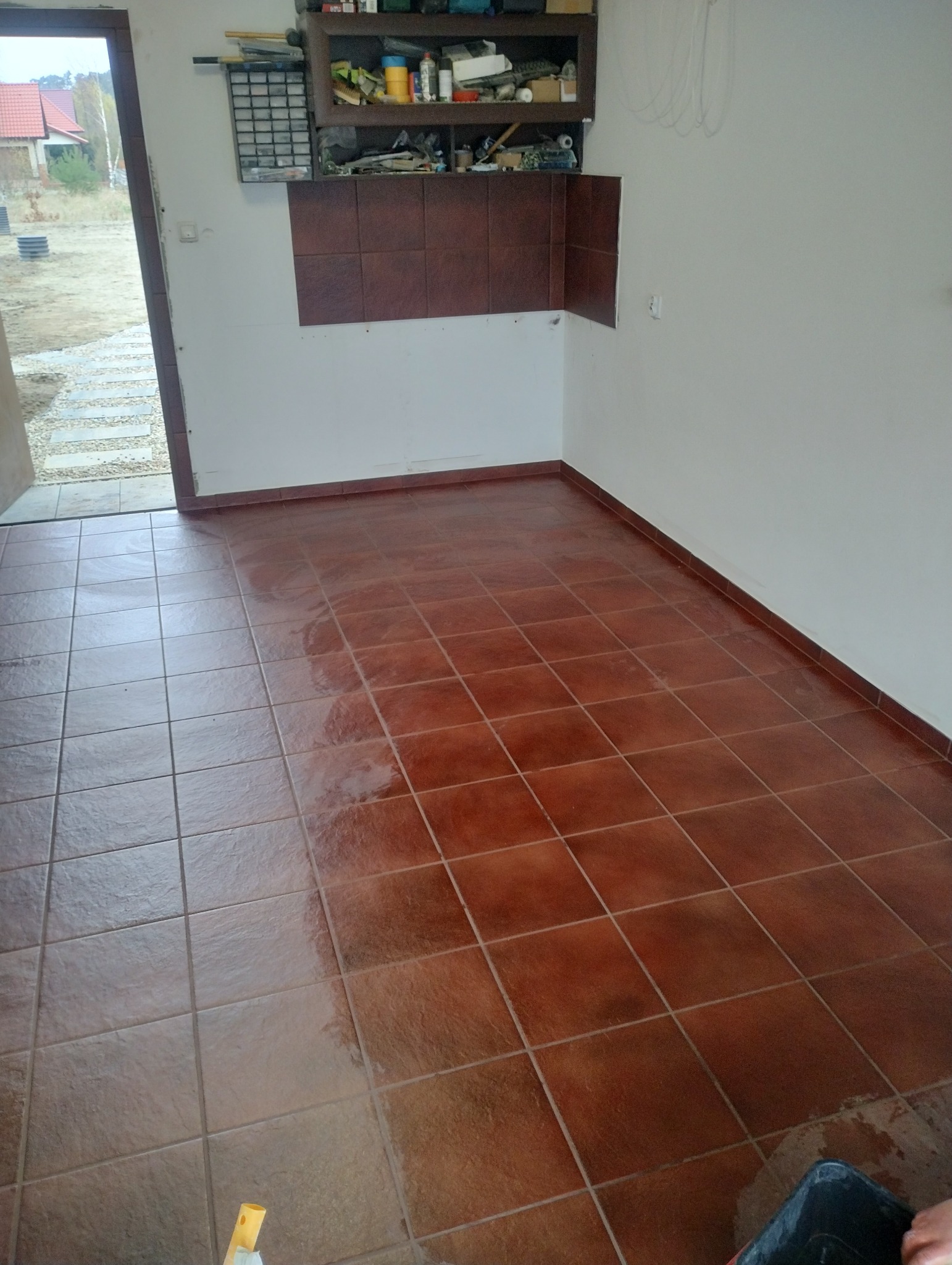 Brązowe płytki podłogowe ułożone w kwadraty, częściowo umyte, z widocznym fragmentem wiadra i mopem w rogu, ściany białe, w tle widok na ogród.