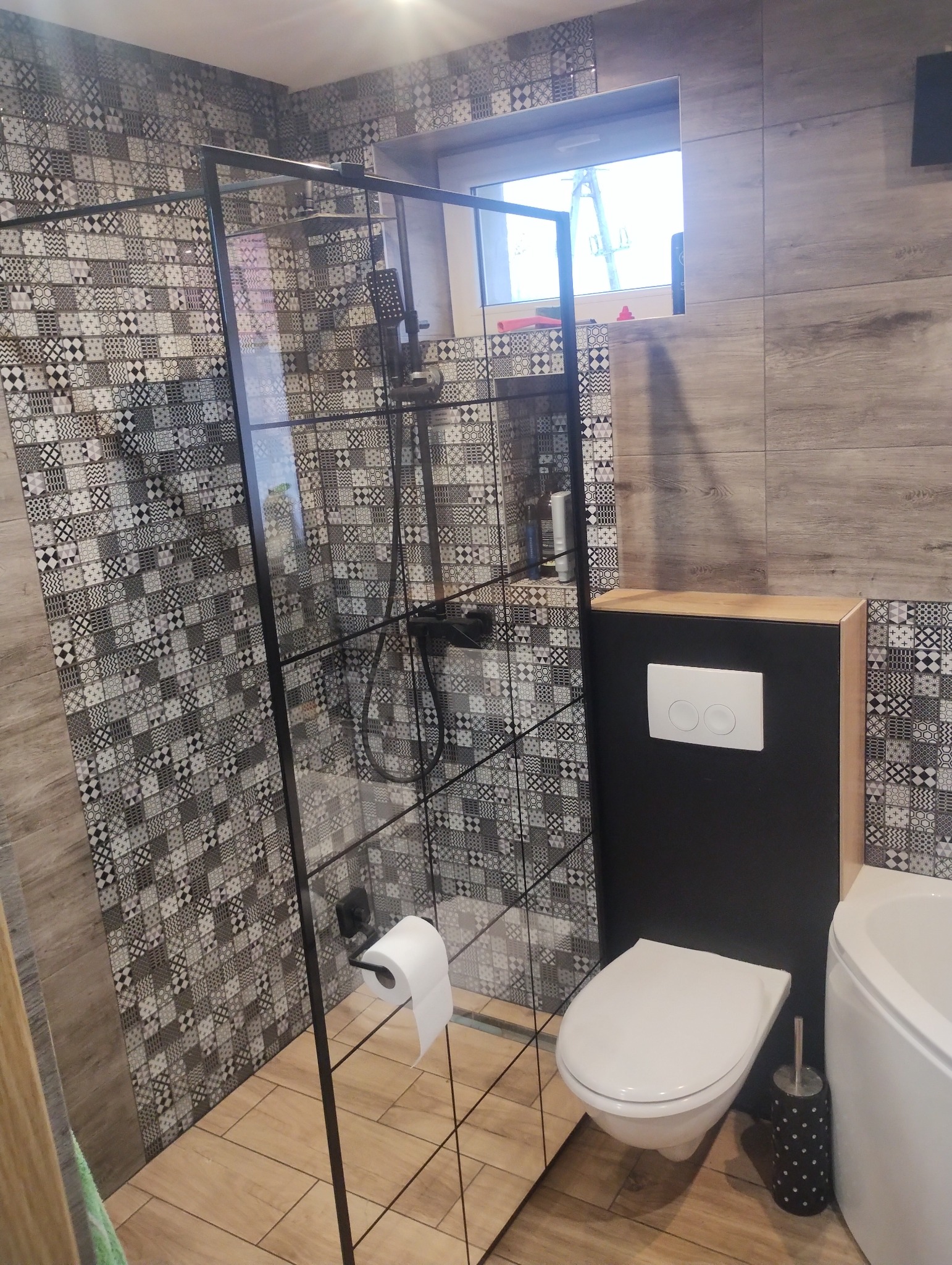 Nowoczesna łazienka z kabiną prysznicową typu walk-in z czarnymi profilami, mozaiką na ścianach i drewnopodobnymi płytkami podłogowymi oraz wiszącą toaletą.