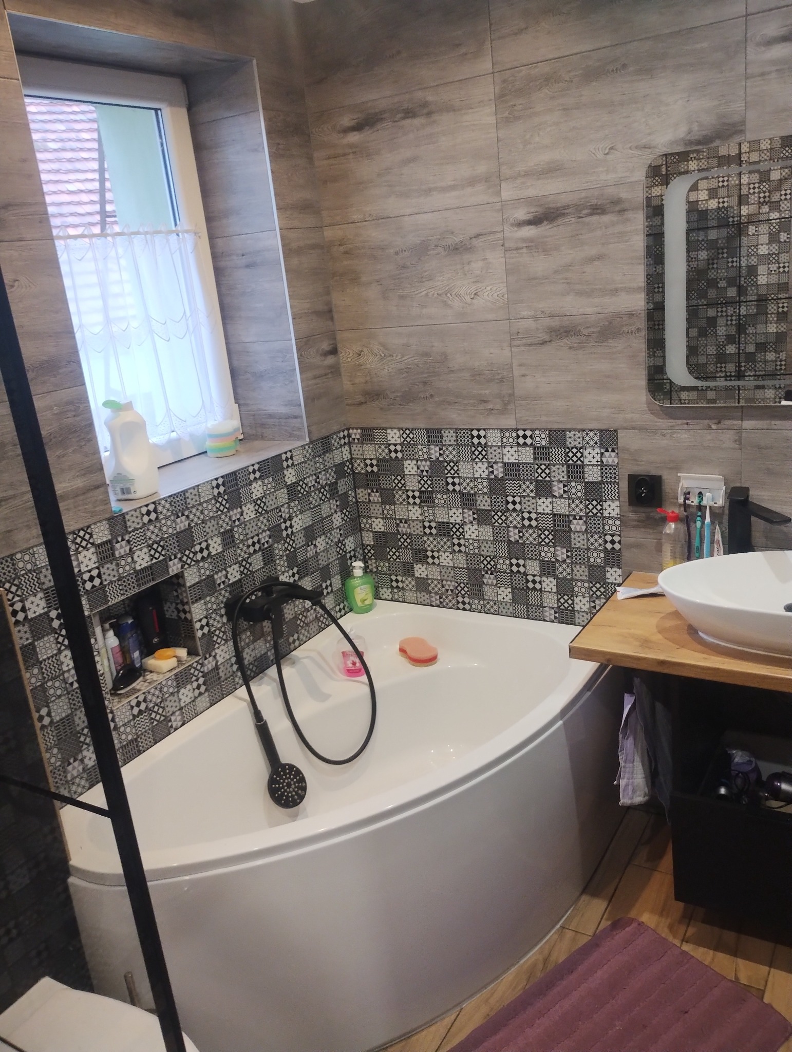 Nowoczesna łazienka z narożną wanną, mozaikową ścianą nad wanną, szarą płytką imitującą drewno na ścianach i podłodze, czarną armaturą i umywalką nablatową na drewnianym blacie.