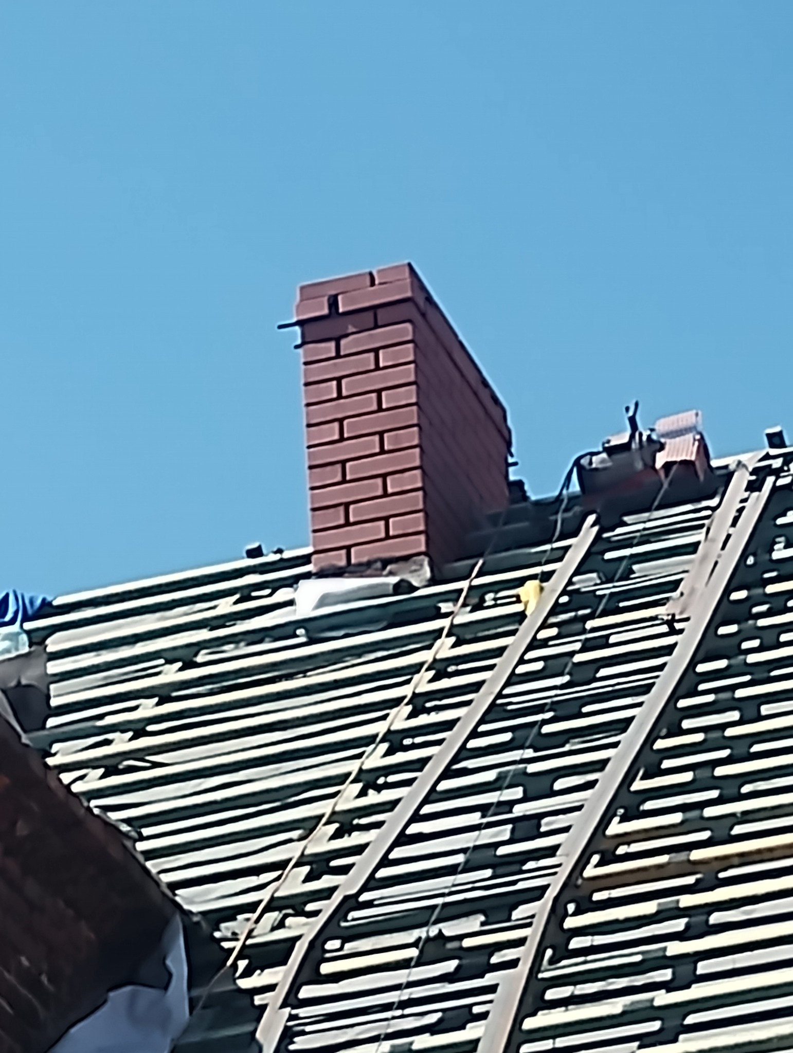 Widok na wznoszący się komin z czerwonej cegły na tle błękitnego nieba, umieszczony na konstrukcji dachu z widocznymi łatami i elementami mocującymi, sugerujący prace dekarskie w toku.