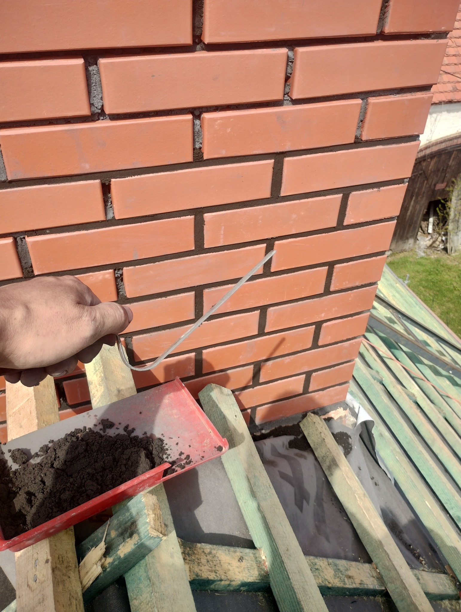 Murowany komin z cegły klinkierowej na dachu, z widoczną ręką murarza, który używa drutu do czyszczenia spoin, obok leży taca z zaprawą.