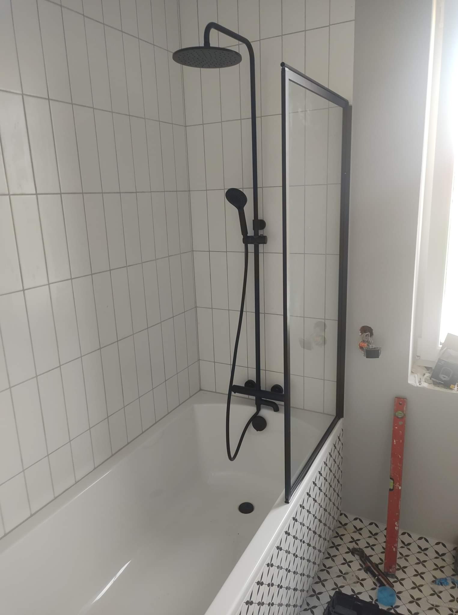 Biała wanna z czarną armaturą prysznicową i szklanym parawanem, białe płytki ścienne i wzorzyste płytki podłogowe, widoczny poziomica i niedokończone gniazdko elektryczne.