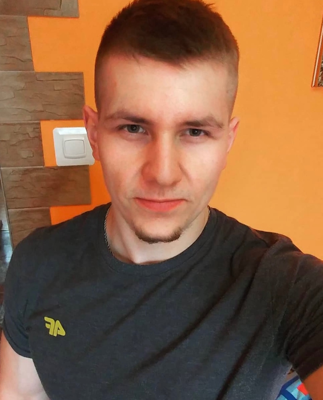 Portret młodego mężczyzny z krótkimi włosami i delikatnym zarostem, ubrany w szarą koszulkę z małym, żółtym logo na tle pomarańczowej ściany z fragmentem dekoracyjnej okładziny.