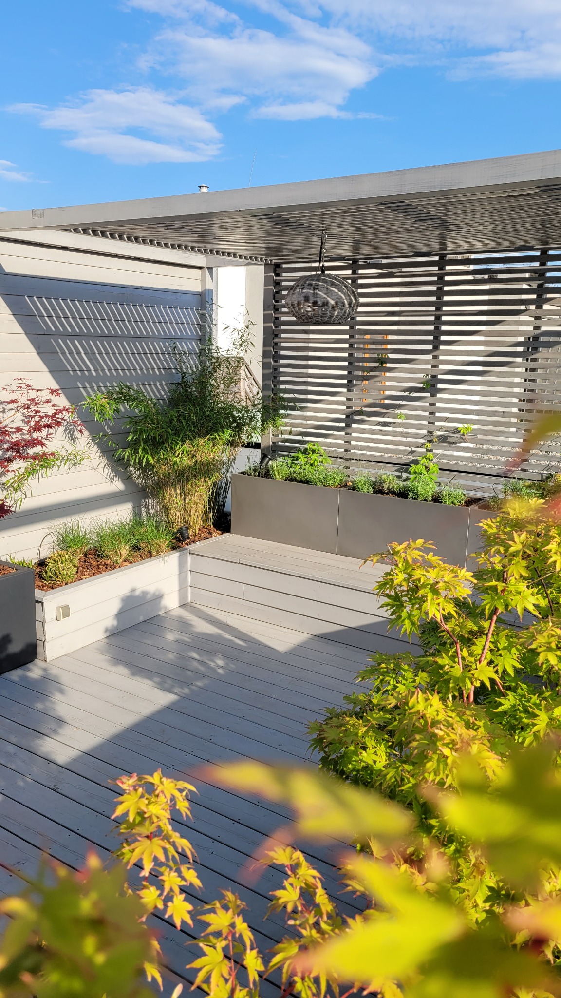 Nowoczesny taras na dachu z drewnianą podłogą, pergolą, metalowymi donicami z zielenią i bambusem, oświetlony wiszącą lampą, w słoneczny dzień.