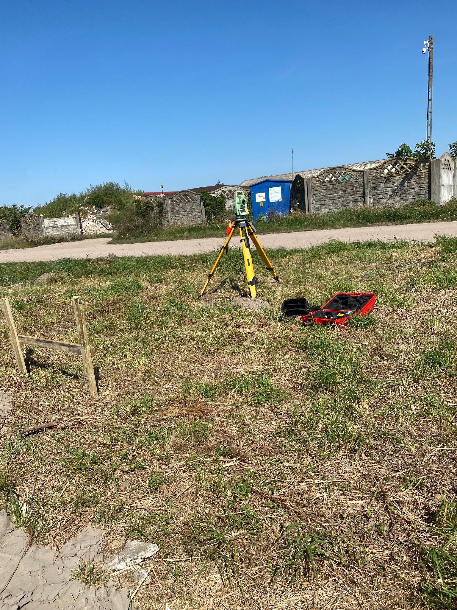 Żółty teodolit ustawiony na trójnogu na trawiastym terenie, obok otwarta czerwona walizka z akcesoriami, w tle niebieski kontener i szary płot.