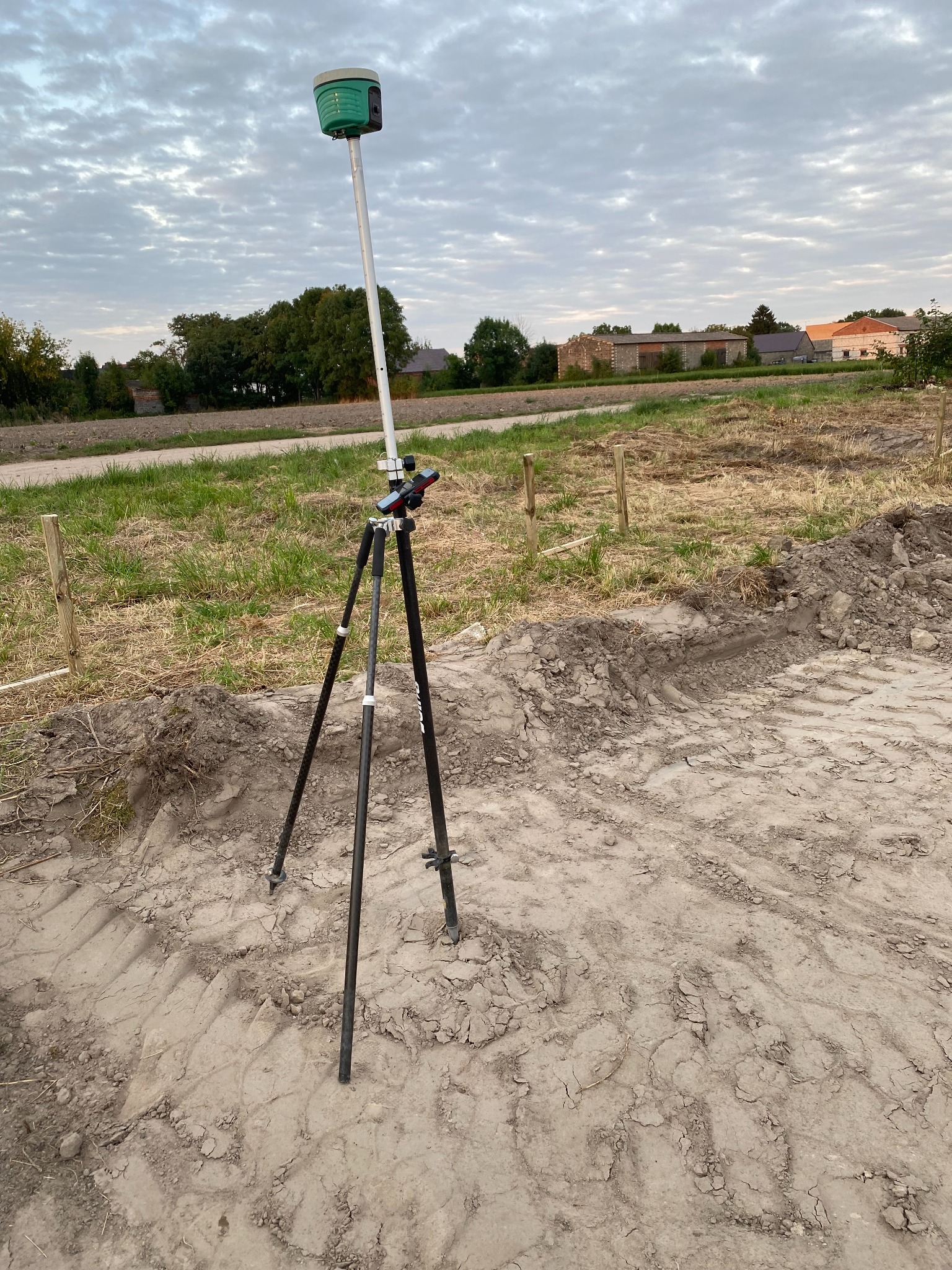 Zielony odbiornik GNSS na czarnym statywie, ustawiony na nierównym, piaszczystym terenie z widocznymi śladami opon, w tle łąka i budynki pod pochmurnym niebem.
