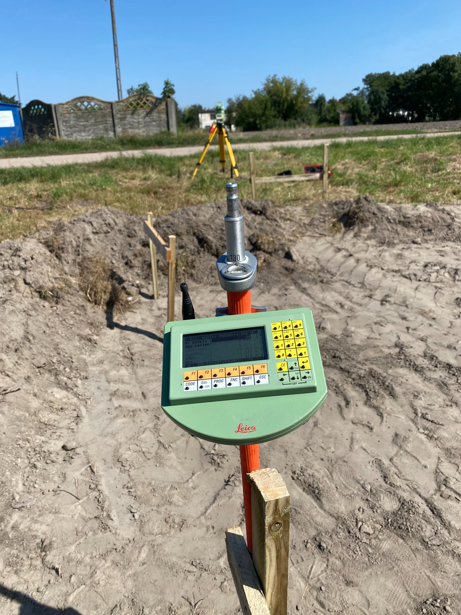 Zielony tachimetr Leica z wyświetlaczem i klawiaturą, ustawiony na tyczce geodezyjnej, w tle wytyczony teren budowy z palikami i widocznym drugim tachimetrem na statywie.