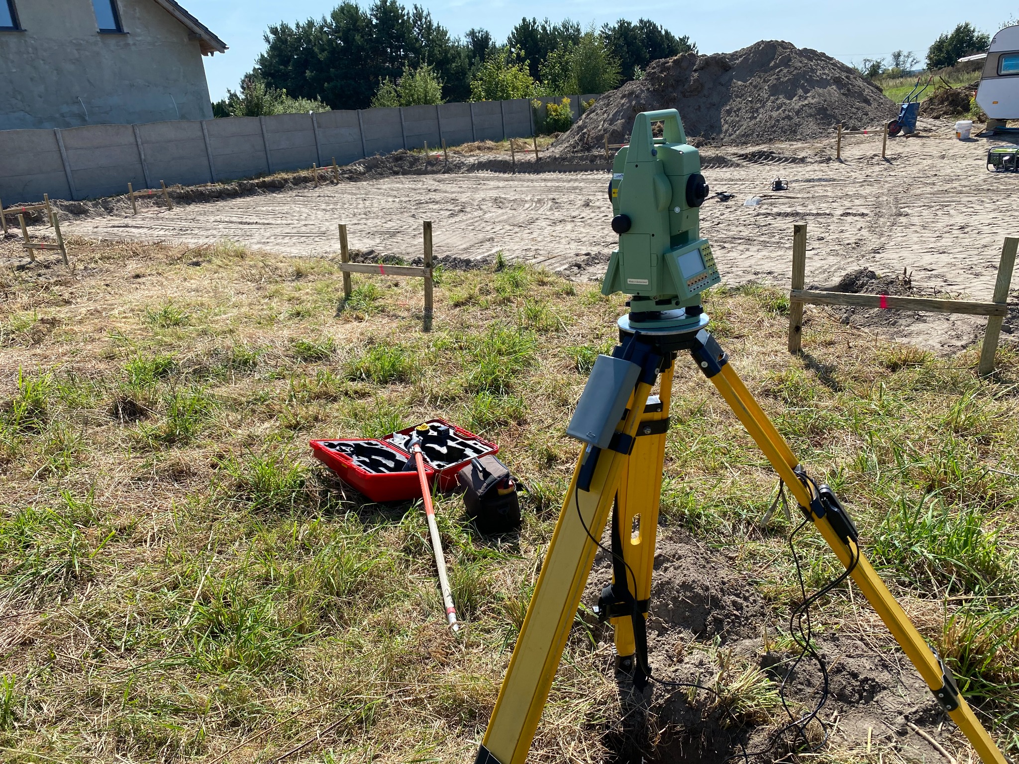 Zielony teodolit na żółtym statywie, skrzynka z akcesoriami pomiarowymi obok, w tle wytyczony teren pod budowę z widocznymi palikami i ogrodzeniem.