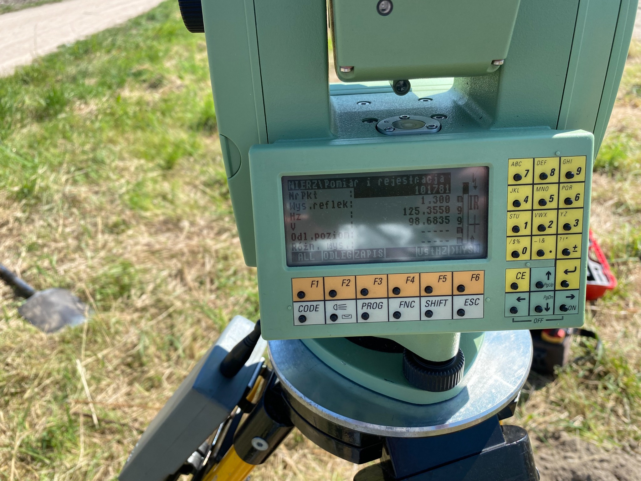 Zielony teodolit z wyświetlaczem pokazującym dane pomiarowe, ustawiony na statywie na trawiastym terenie, obok widoczna łopata.