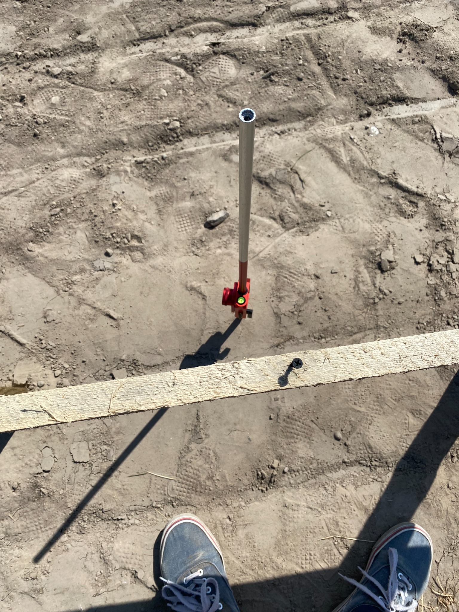 Niwelator laserowy ustawiony na gruncie obok drewnianej deski z wkręconą śrubą, widok z góry na stopy w trampkach.