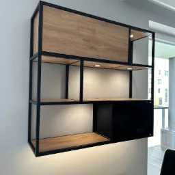 Półka wisząca z drewnianymi i czarnymi elementami, oświetleniem punktowym LED, zamontowana na szarej ścianie nad grzejnikiem.