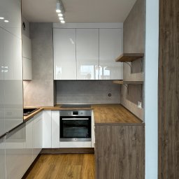 Salon Meblowy Bonkowscy - Nowoczesna kuchnia z białymi, lakierowanymi frontami i drewnianymi blatami. Widoczne oświetlenie punktowe i półki z metalowymi wspornikami. Realizacja w jasnej tonacji.