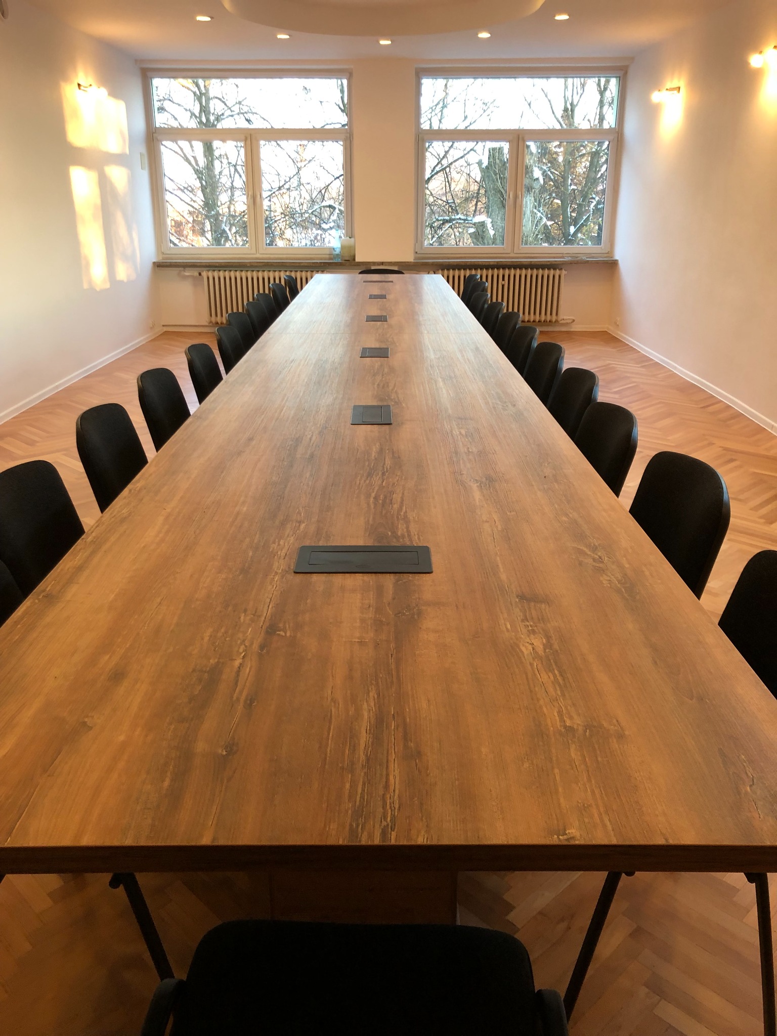 Długi, drewniany blat stołu konferencyjnego z wbudowanymi gniazdami, otoczony czarnymi krzesłami w jasnym pomieszczeniu z parkietem i dużymi oknami.