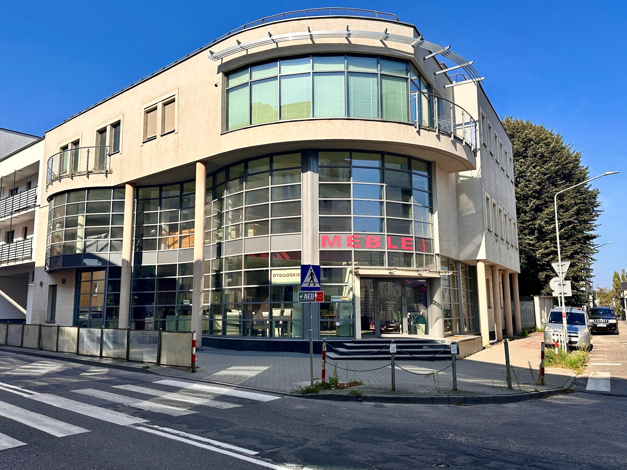 Fasada dwupiętrowego salonu meblowego z dużymi oknami wystawowymi, widok z ulicy z przejściem dla pieszych.